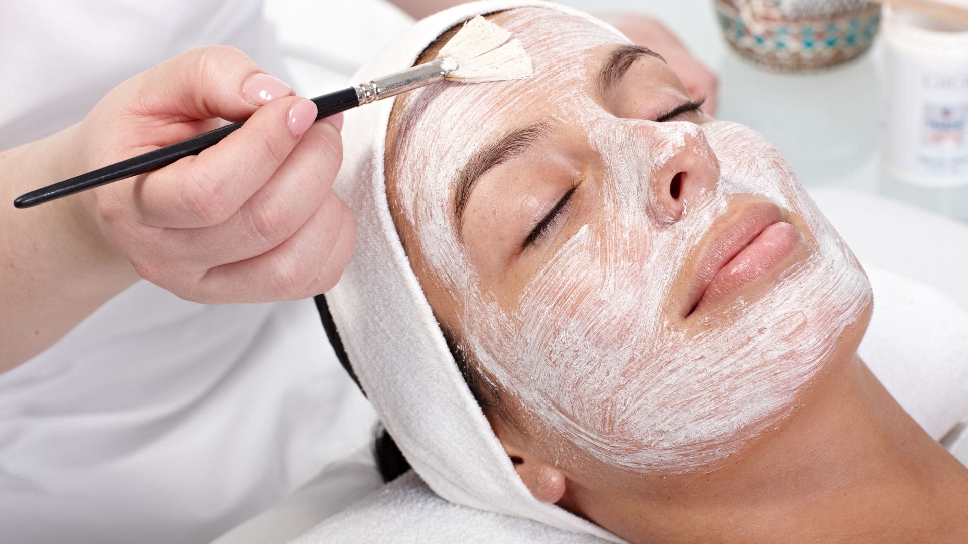 Kosmetikerin trägt eine Gesichtsmaske mit einem Pinsel auf das Gesicht einer Kundin auf. Behandlungsszene für Gesichtsbehandlungen.